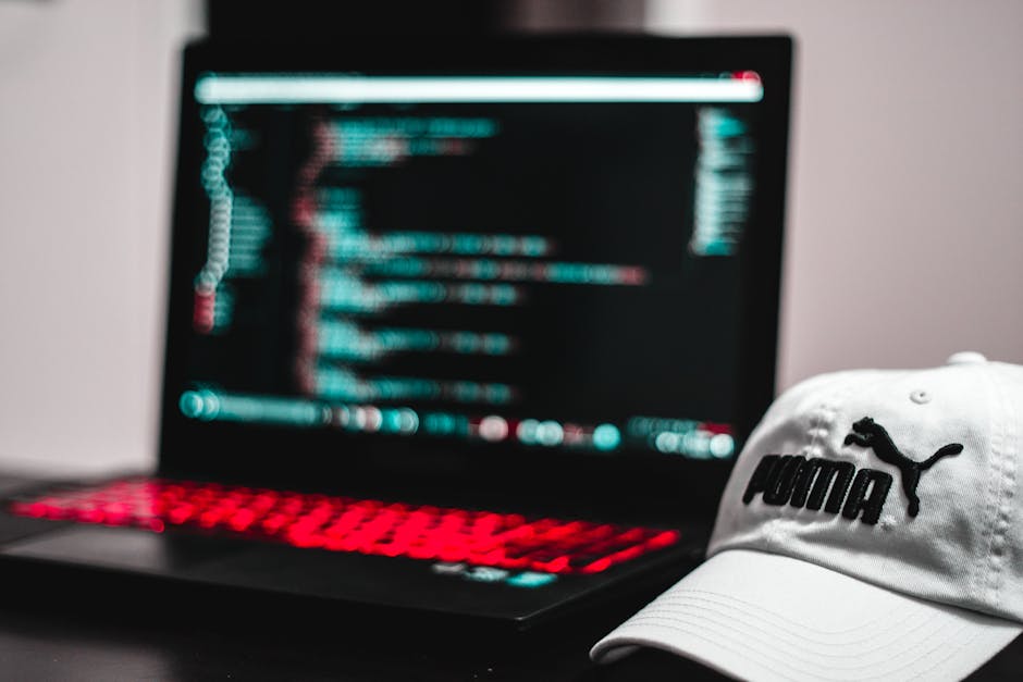 A laptop with a black keyboard illuminated by red backlighting displaying lines of colorful code on the screen, positioned on a dark surface, with a white Puma cap featuring the black Puma logo placed